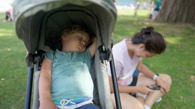 Child napping laying inside stroller with mother looking at phone sitting at park during summer sunny day. Little boy with eyes closed sleeping in the afternoon