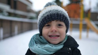 Kış mevsiminde karla kaplı mutlu bir çocuk portresi. Dışarıda kameraya gülümseyen bereli atkı ve palto giyen küçük bir çocuk.
