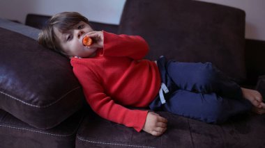 Kanepede uzanmış havuç yiyen küçük bir çocuk. Çocuk kanepede oturup kameradan çizgi film izliyor.