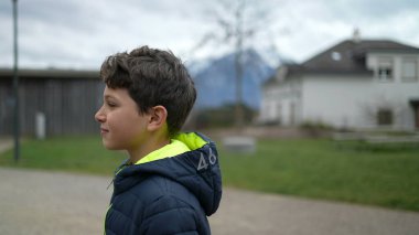 Parkta ileri doğru yürüyen dalgın bir genç çocuk. Üzerinde kışlık giysiler olan düşünceli bir çocuk şehir parkında yürüyor.