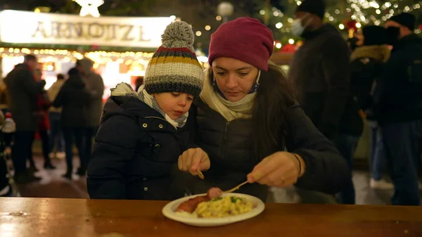 Mother and child eating food in winter Christmas market in Europe. Mom feeding little boy food seated at table during festivities
