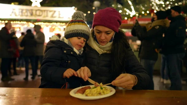 Mother and child eating food in winter Christmas market in Europe. Mom feeding little boy food seated at table during festivities