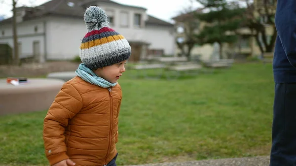 One cute little boy wearing beanie and jacket during winter season standing outdoors at park observing friends play with toy. Tracking shot