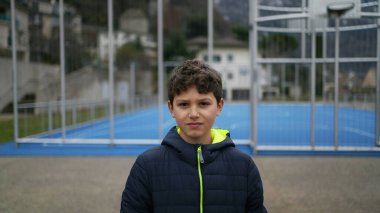 Contemplative young boy standing outside at city park with serious expression. Thoughtful preteen kid wearing winter clothes stands outdoors. Portrait face