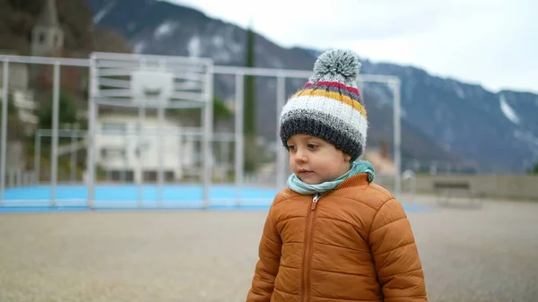 One cute little boy wearing beanie and jacket during winter season standing outdoors at park observing friends play with toy. Tracking shot