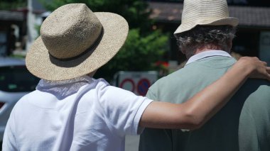 Ergen bir çocuğun kollarıyla büyükbabayı kucaklaması. İki kuşak aile ferdi kucaklaşarak dışarıda yürüyorlar. Şapka takıyorlar.