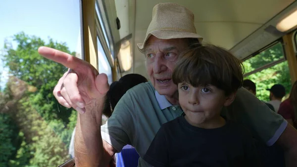 Happy grandfather traveling by train with grandchild together. Senior man pointing at landscape inside railroad wagon in motion. Family on vacation