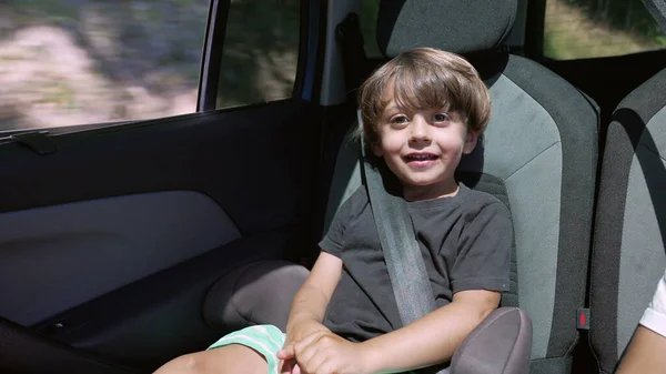 一个快乐的小男孩坐在车后座上对着摄像机笑 有安全带的儿童在路上行走 — 图库照片