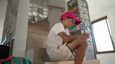 Telefonunu kullanıp ekranlara bakan genç bir çocuk. Evdeki merdivenlerde oturuyor. Akıllı telefon cihazında sosyal medyaya kayan bir genç çocuk