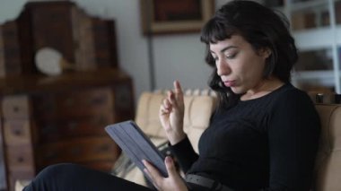 Latin kökenli genç bir kadın internetten tablet kullanıyor. Zarif oturma odasında oturuyor.