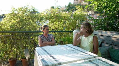 Anne, terasta yetişkin kızıyla konuşuyor. Yaz günü dışarıda sohbet eden iki kişi.
