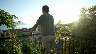 Düşünceli yaşlı bir adam dışarıda dikilip evden manzaraya bakıyor. Emekliliğin tadını çıkaran yaşlı bir erkeğin arkası manzaralı.