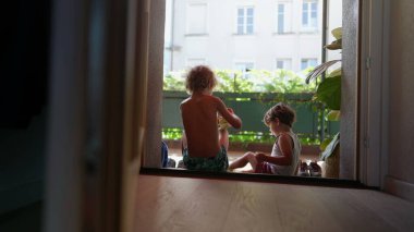 Çocuklar kapının önünde oturuyor. Ayakkabılarını dışarıda giyiyorlar. Büyük kardeş, küçük kardeşe ayakkabıyı kapının önüne koyması için yardım ediyor.