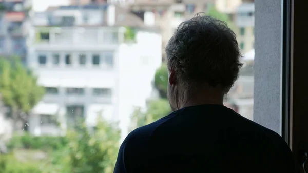 stock image Back of a contemplative senior man looking out window. Pensive older person standing at home staring at view in silhouette