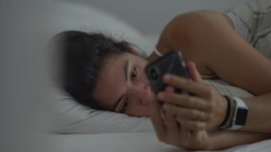 Akıllı telefondan yatakta yatan ciddi bir kadın. Battaniyenin altındaki telefon ekranına bakan bir kadın.