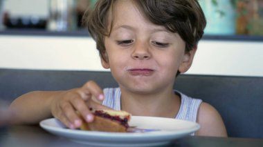 Kahvaltı masasında oturan küçük bir çocuk jöleli ekmeği çiğniyor.