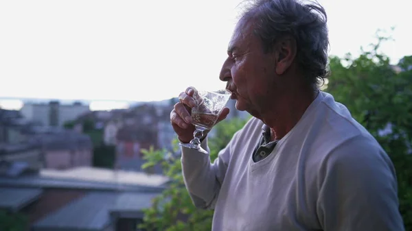 One senior man drinking wine while standing outdoors looking at view. Mature older person enjoying retirement drinks aloholic beverage observing scenic landscape
