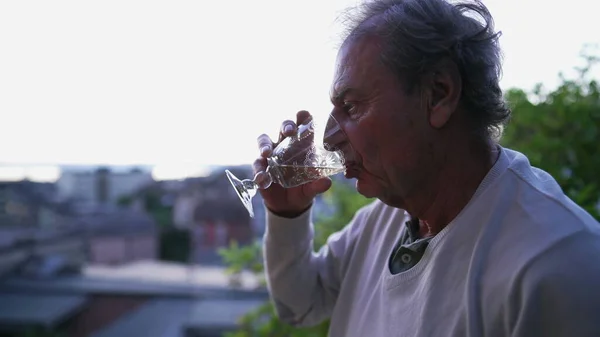 One senior man drinking wine while standing outdoors looking at view. Mature older person enjoying retirement drinks aloholic beverage observing scenic landscape