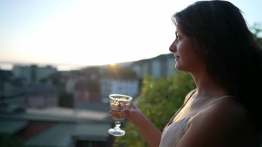 Gün batımında beyaz şarap içen bir kadın. Yaz günü açık havada alkollü içecekten hoşlanan bir kadın manzaraya bakıyor.