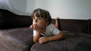 Çocuk TV 'de medyayı izlerken kanepede uzandı. Küçük bir çocuğun portresi ekrandan dışarı bakıyor.