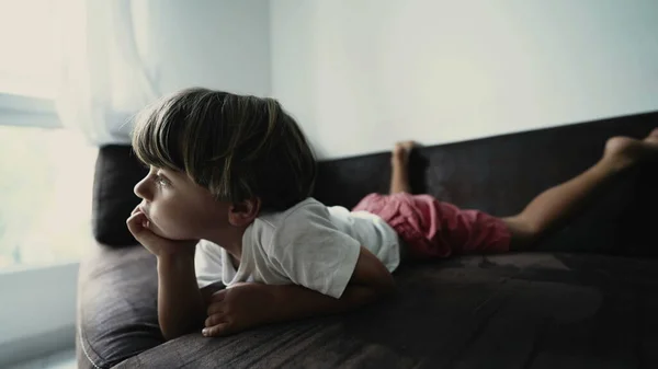 One small boy lying on couch watching cartoons off camera. A relaxed kid laid on sofa