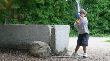 Child getting water from fountain and spraying with foam blaster toy gun outdoors