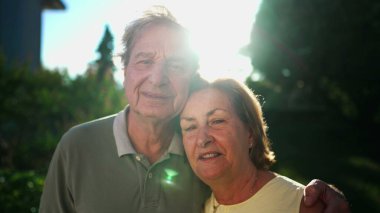 A happy married older couple looking at camera at park with sunlight flare backlight. Close up of senior faces of 70s husband and wife