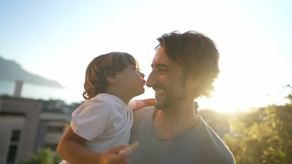 Child son kissing father many times. Parent holding kid standing outdoors in sunset time. Love and fatherhood concept