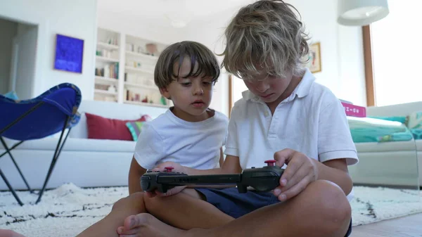 Children looking at video game screen tablet. Younger brother watching sibling play games on joystick console. Kids playing with technological device at home living room