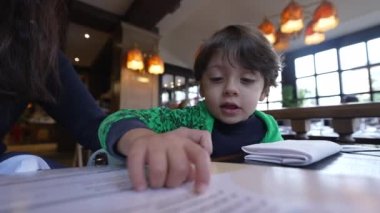 Küçük çocuk restoran menüsü okuyor. Çocuk yemek seçerken masada oturuyor.