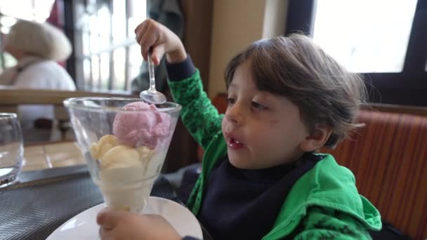 小さな男の子がアイスクリームデザートを食べています 子供はレストランに座ってスプーンでおいしいイチゴとバニラアイスを食べます — ストック動画