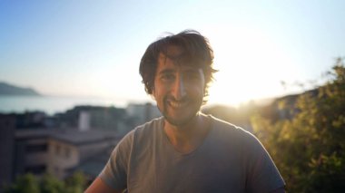Portrait of a happy man in 30s standing outdoors in sunset time overlooking landscape in background. Close up face male person