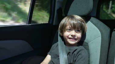 Arabasının arka koltuğunda oturan mutlu, küçük bir çocuk. Emniyet kemeri takılı çocuk hareket halinde.