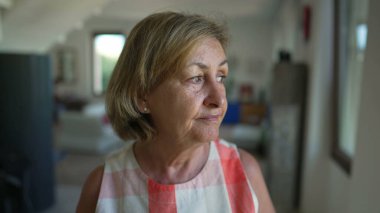 Portrait of a contemplative senior woman standing indoors at home living room looking out window. Pensive expression