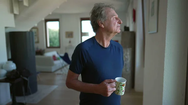 Stock image One thoughtful senior man standing at home living room looking out window with pensive expression. 70s male older mature person holding cup of coffee in morning