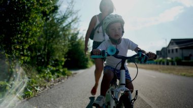 Anne oğluna bisiklet yolunun dışında bisiklet sürmeyi öğretiyor. Kask takan başarılı bir çocuk bisiklete binip pedal çeviriyor..