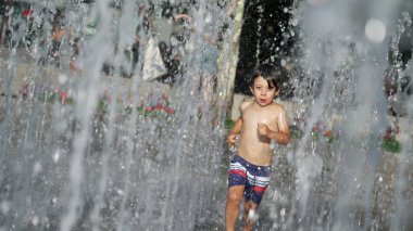 Küçük çocuk yaz günü şehir parkında su jetlerinde koşuyor. Küçük çocuk serinletici sularda koşar