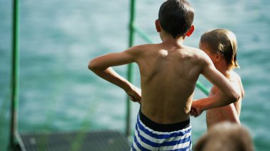 Göl kenarında duran iki ıslak çocuğun arkasında. Mayo ayarlayan genç bir çocuk. Çocuklar yaz tatilinin tadını çıkarıyorlar