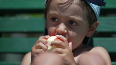 Küçük çocuk yaz günü dışarıda elma meyvesinden bir ısırık alıyor. Düşünceli şirin küçük çocuk dışarıda güneş ışığında sağlıklı şeyler yiyor.