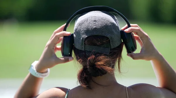 stock image Back of person head putting headphones sitting outside at park bench. Young woman listening to music song podcast or audiobook outdoors