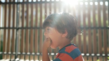 Evde balkonda oturan küçük bir çocuk, elinde güneş ışığıyla mısır gevreği yiyor. Çocuk pijama giyerek yemek yiyor.