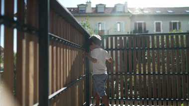 Oyuncu bir çocuk apartmanın balkonunda dikilip ikinci kattaki ahşap çit korumasından bakıyor.