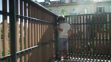 Oyuncu bir çocuk apartmanın balkonunda dikilip ikinci kattaki ahşap çit korumasından bakıyor.