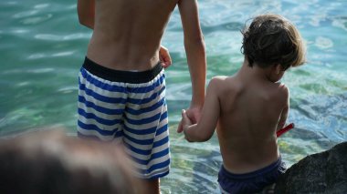 İki kardeş göl kenarında el ele tutuşup mayo giymişler. Aile yaz tatilinin tadını çıkarıyor. Kardeşler küçük kardeşe yardım eder.