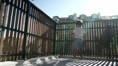 Balkon çitinin yanında duran küçük bir çocuk ikinci kattan dışarı bakıyor. Tahta parmaklıklarda bekleyen çocuk dışarı bakıyor.