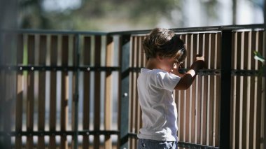 Düşünceli küçük bir çocuk ikinci kattaki apartman evinin ahşap çitlerine tutunarak balkonda duruyor. Düşünceli çocuk dışarı bakıyor