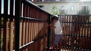 Çocuk apartmanın balkonunda durmuş dışarıdaki ahşap çitlerden bakıyor. İkinci kattaki terastan bakan küçük bir çocuk.