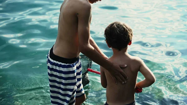 十多岁的哥哥牵着年幼的兄弟姐妹的手站在湖边 两个穿着泳裤的孩子在一起玩 表妹们在户外玩得很开心 — 图库照片