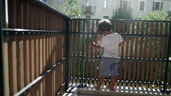 stock image One playful little boy standing at apartment balcony in sunlight. Child holding on terrace fence with lens flare. Kid looking outside through wooden porch