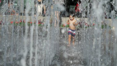 Küçük çocuk yaz günü şehir parkında su jetlerinde koşuyor. Küçük çocuk ferahlatıcı suda koşuyor yavaş çekimde sıçrıyor